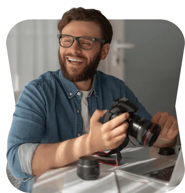 Smiling photographer holding a camera at his desk next to a laptop